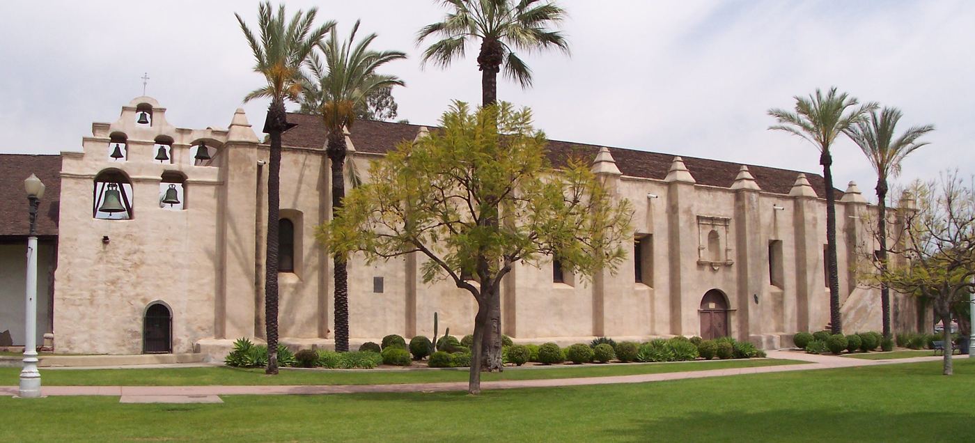 The long whitewashed adobe side wall of Mission San Gabriel, with its bell wall at the left, palm trees and a green lawn