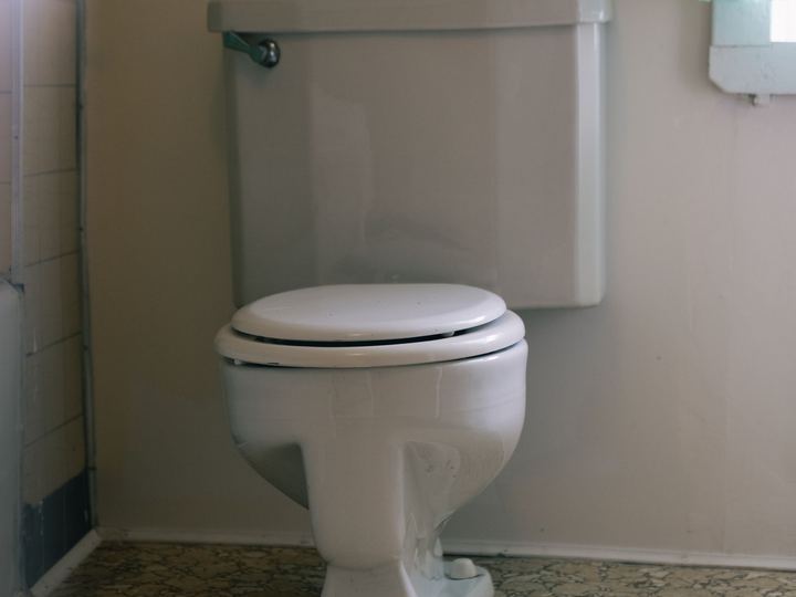 Older pale gray two-piece toilet with a chrome handle on a patterned vinyl floor