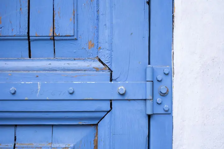 Fragment of weathered wooden door of blue color with metal hinge near white wall